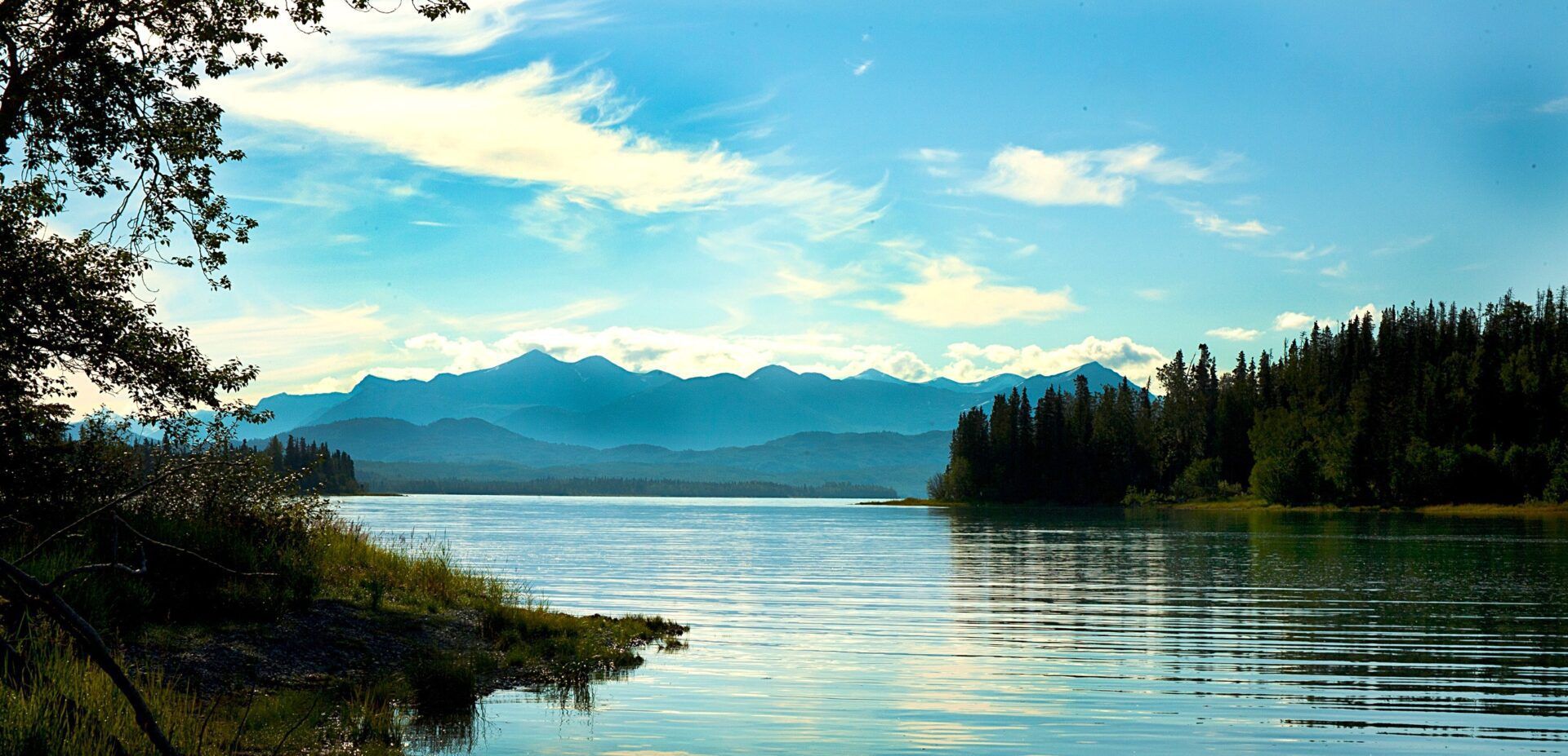 A serene landscape featuring a calm, blue lake reflecting mountains in the distance, framed by dark trees on the shore.