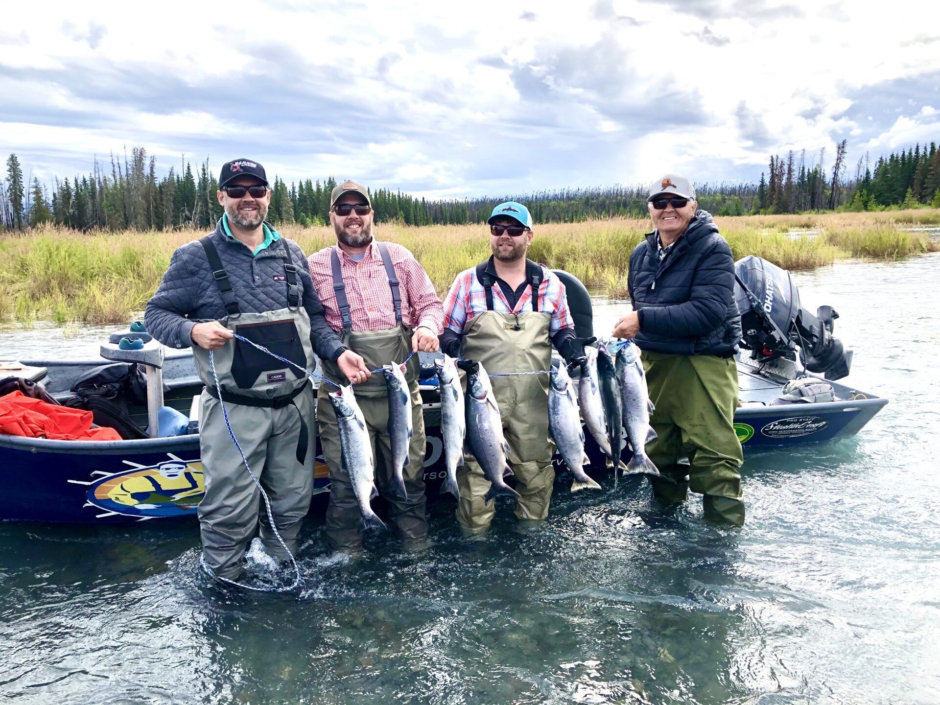 Four people in fishing gear stand in a shallow river next to a boat, holding up several caught fish.