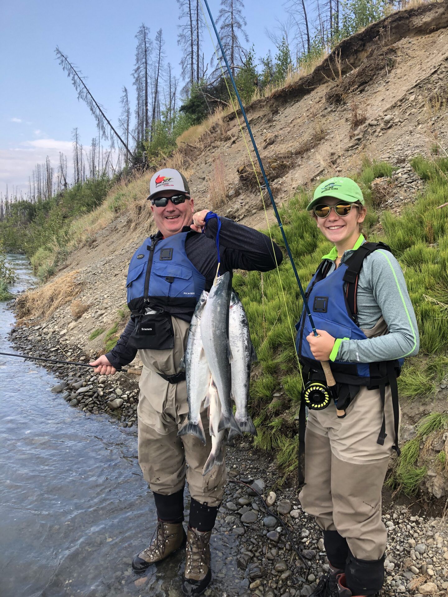 Two people in waders and life jackets stand on a riverbank holding caught fish while holding fishing poles.
