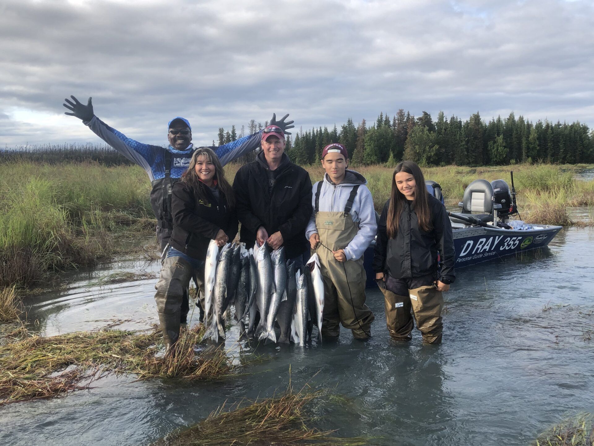 A group stands in shallow water holding a string of fish, with a boat docked behind them in a grassy, wooded landscape.
