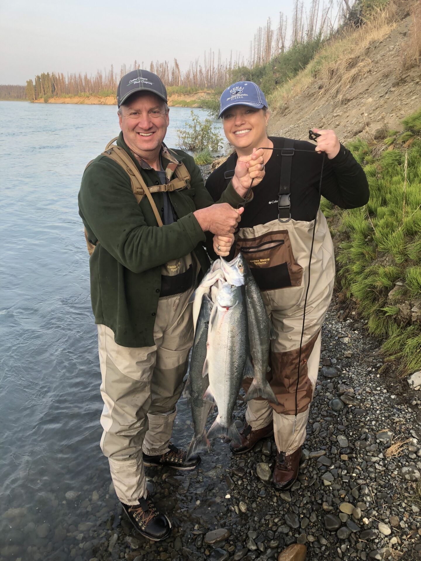 Two people in fishing gear stand on a riverbank holding two caught fish, smiling at the camera.