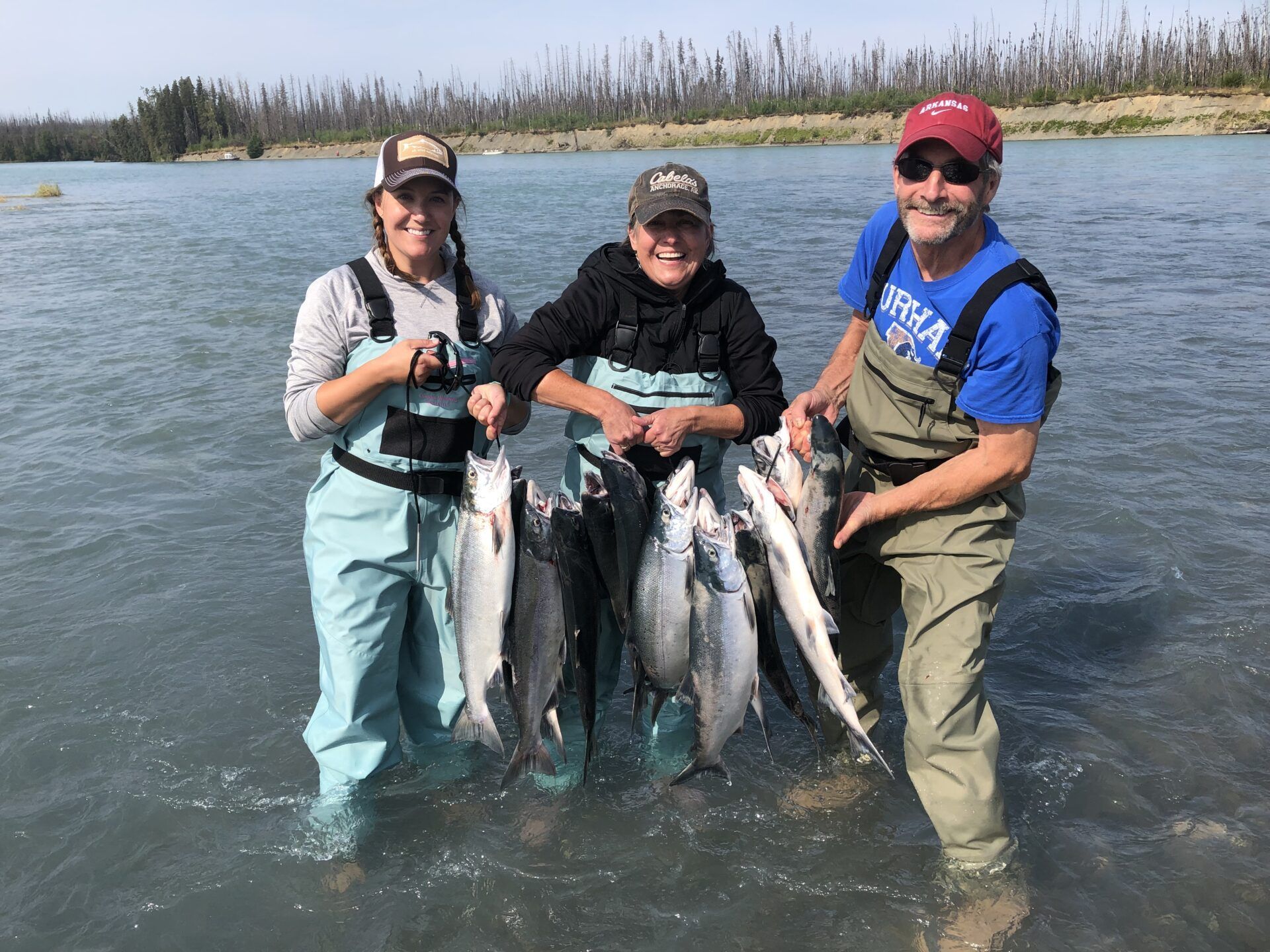 Three people in waders standing in a river holding several caught salmon under a clear blue sky.