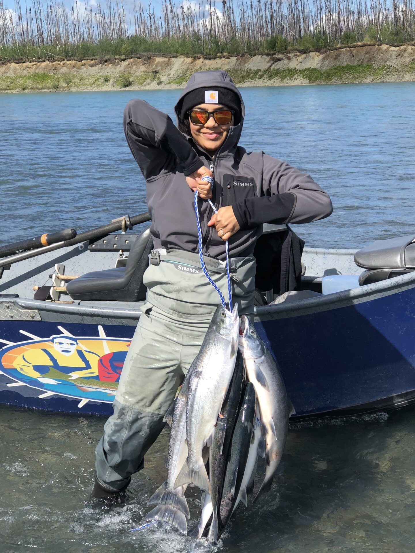 A person in fishing gear and sunglasses stands in a river, holding a stringer of freshly caught salmon by a boat.
