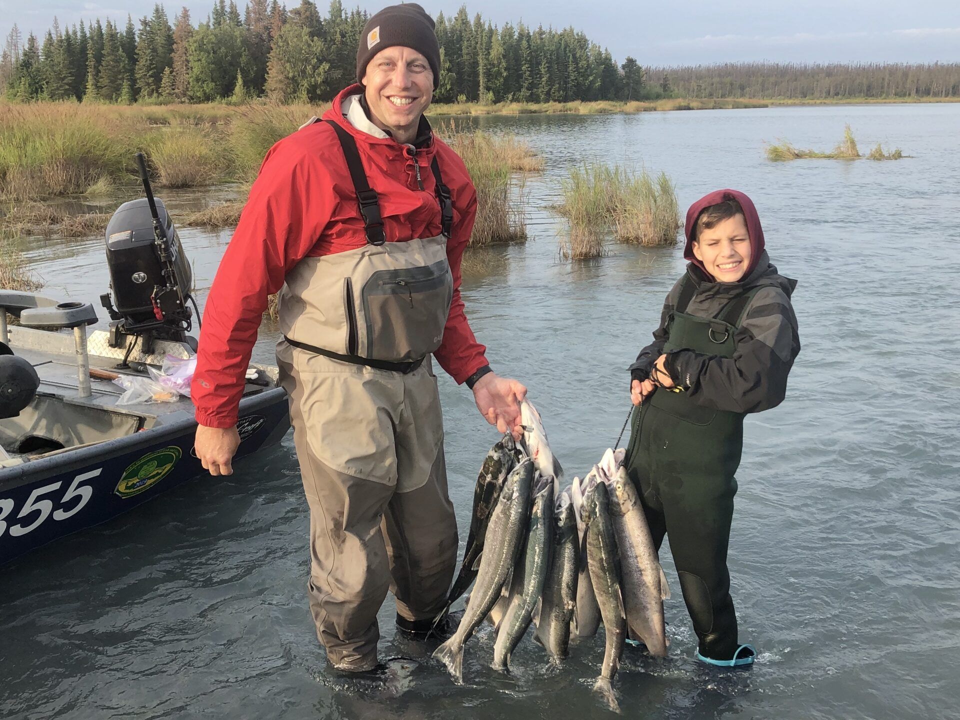 A smiling adult and child in waders stand in a river next to a boat, holding a large stringer of freshly caught fish.