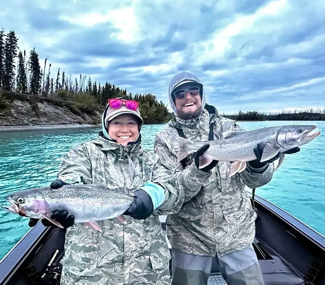 Two people in camouflage fishing gear hold trout while smiling on a boat in a river with a cloudy sky.