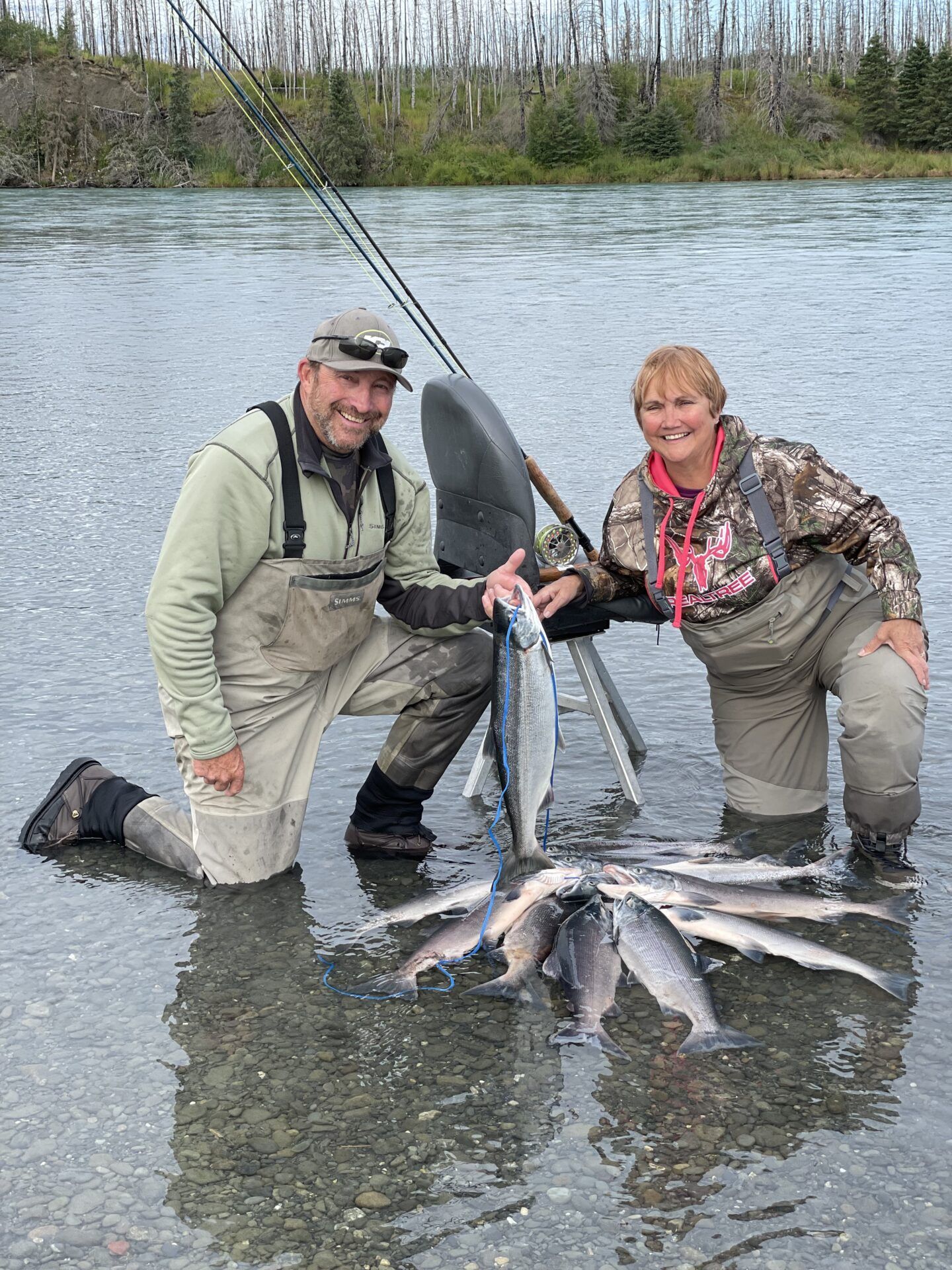 A man and a woman in fishing gear kneel in a river with their catch of several salmon.