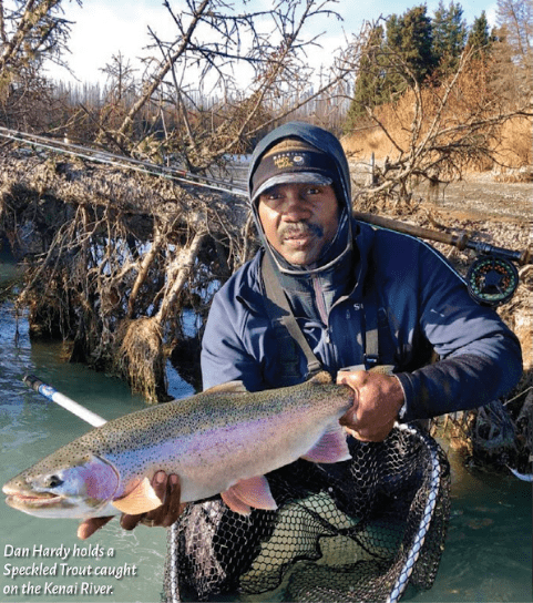 A person wearing a hooded jacket and cap holds a rainbow trout caught in the Kenai River.