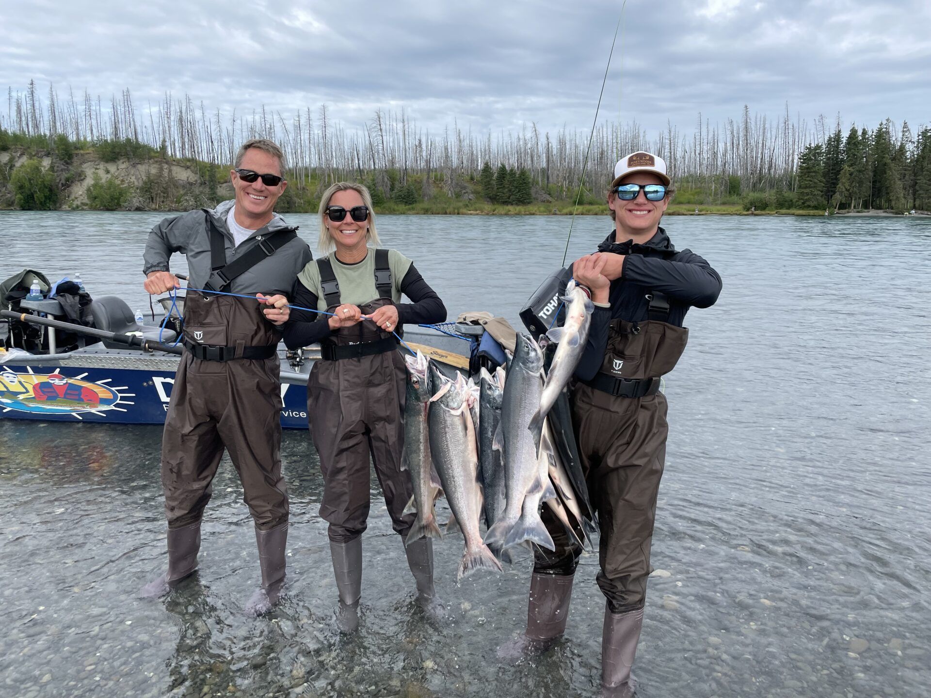 Three people in waders stand in a river holding several caught fish, with a fishing boat parked behind them.