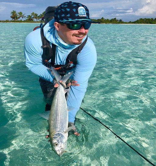 A person in blue sun-protective clothing stands in clear, shallow water holding a large fish, wearing a hat and sunglasses.