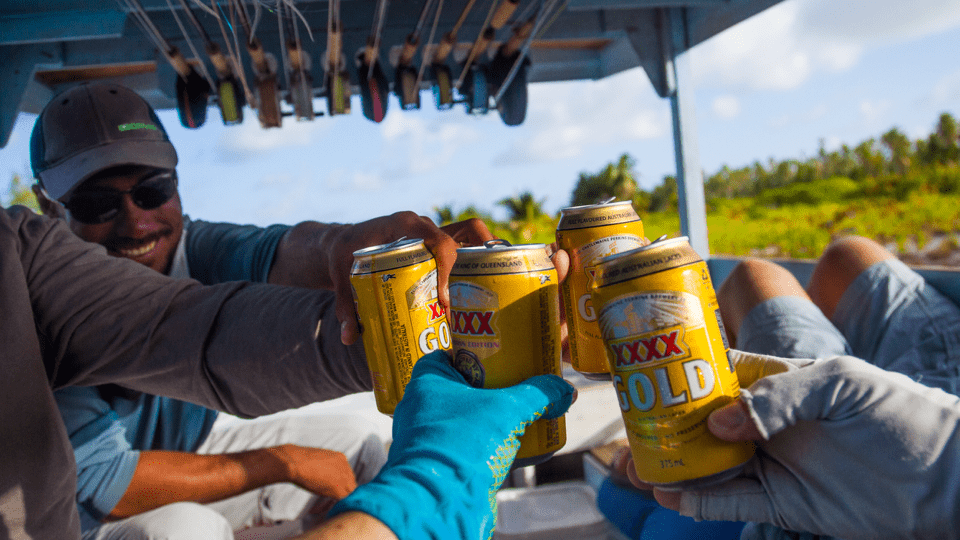 Three people on a boat toast with cans of XXXX Gold beer in an outdoor, sunny setting.