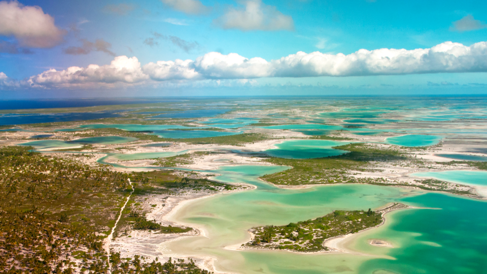 Aerial view of shallow turquoise waters, sandy flats, and small green islands under a blue sky with soft white clouds.
