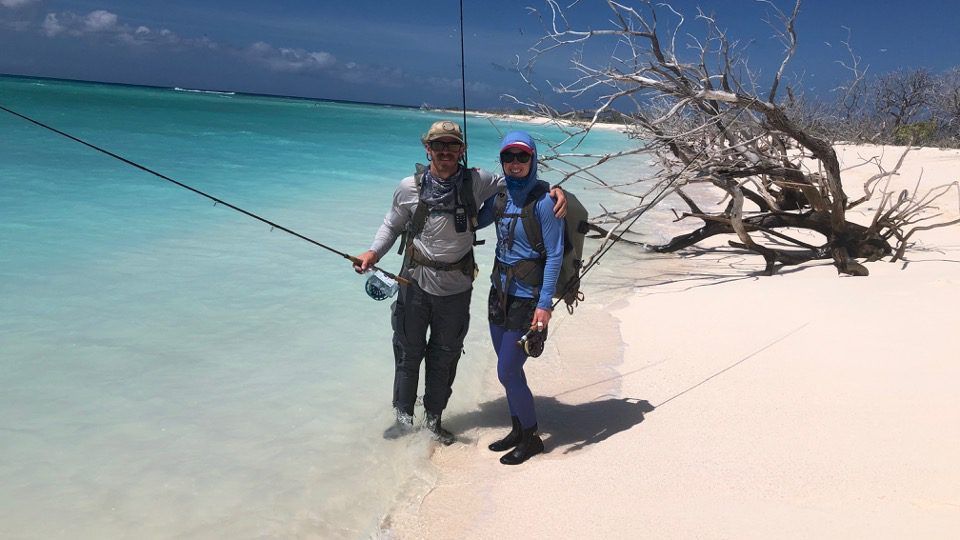 Two people in outdoor gear and backpacks stand on a white sand beach by turquoise water, holding fishing rods.