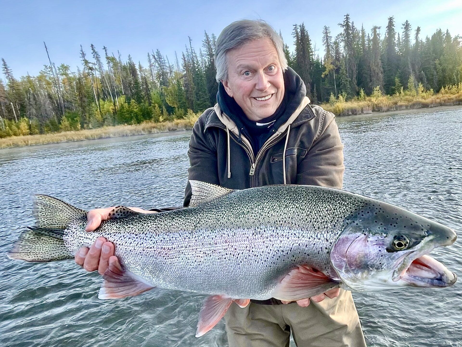 A person smiles while holding a large rainbow trout in a river with a forest background.