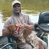 A person wearing a hooded jacket and sunglasses sits on a boat, petting a brown spotted dog standing on their lap.