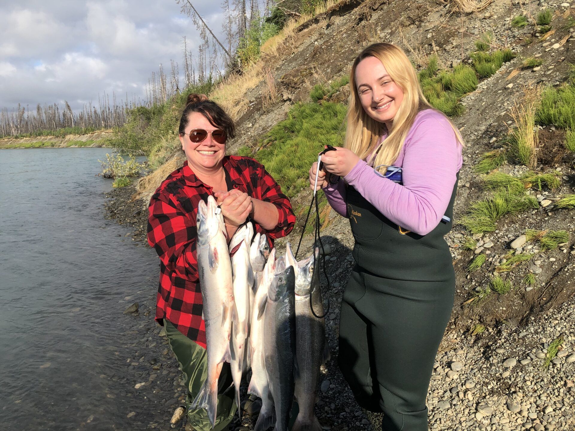 Two people holding a cluster of freshly caught fish while standing on a rocky riverbank.