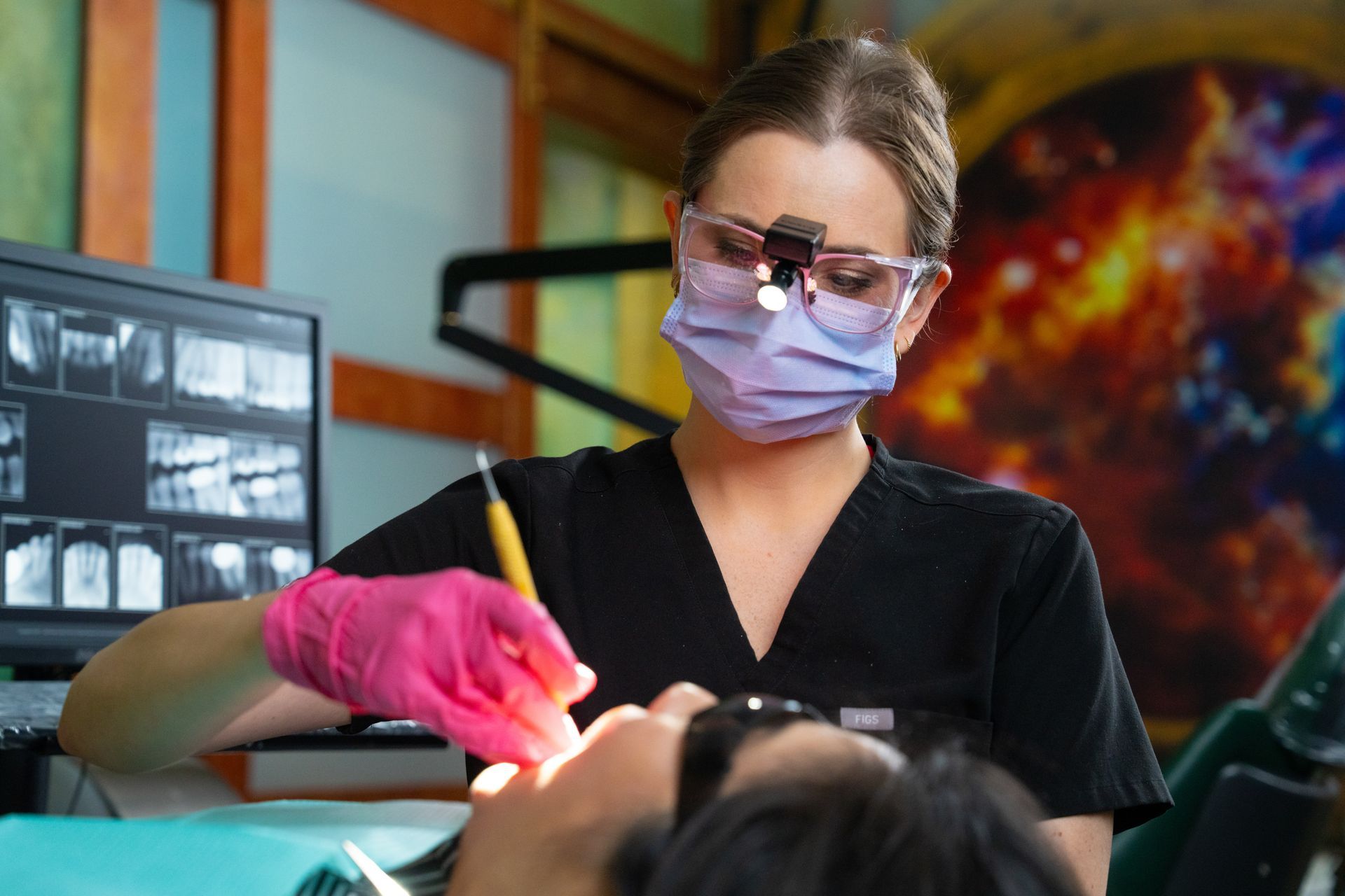 Dentist wearing mask and glasses works on a patient in a dental clinic with monitors and colorful wall art.