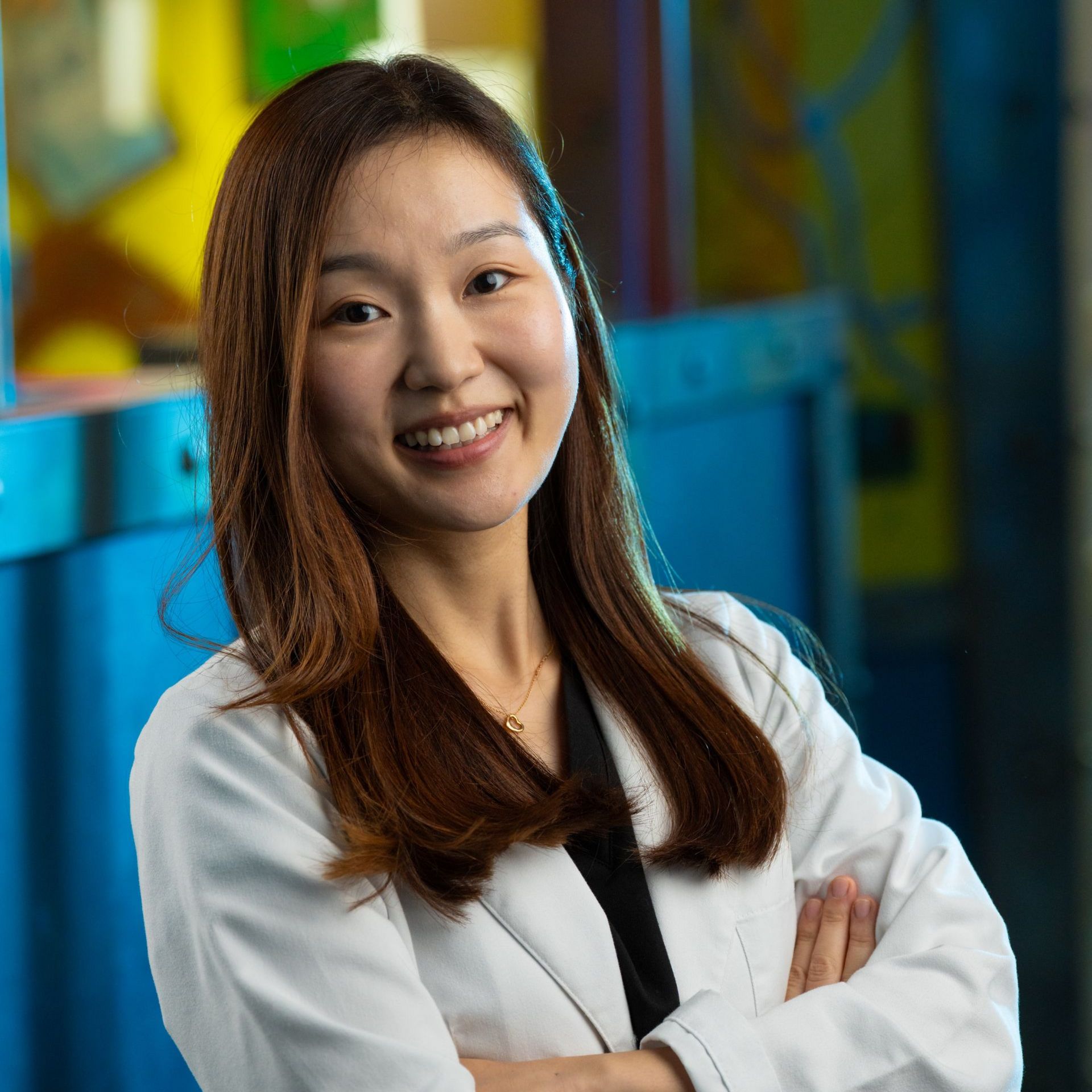 Portrait of a smiling professional in a white lab coat, arms crossed, in a colorful office setting