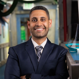Smiling man in a navy suit standing in a modern office hallway.