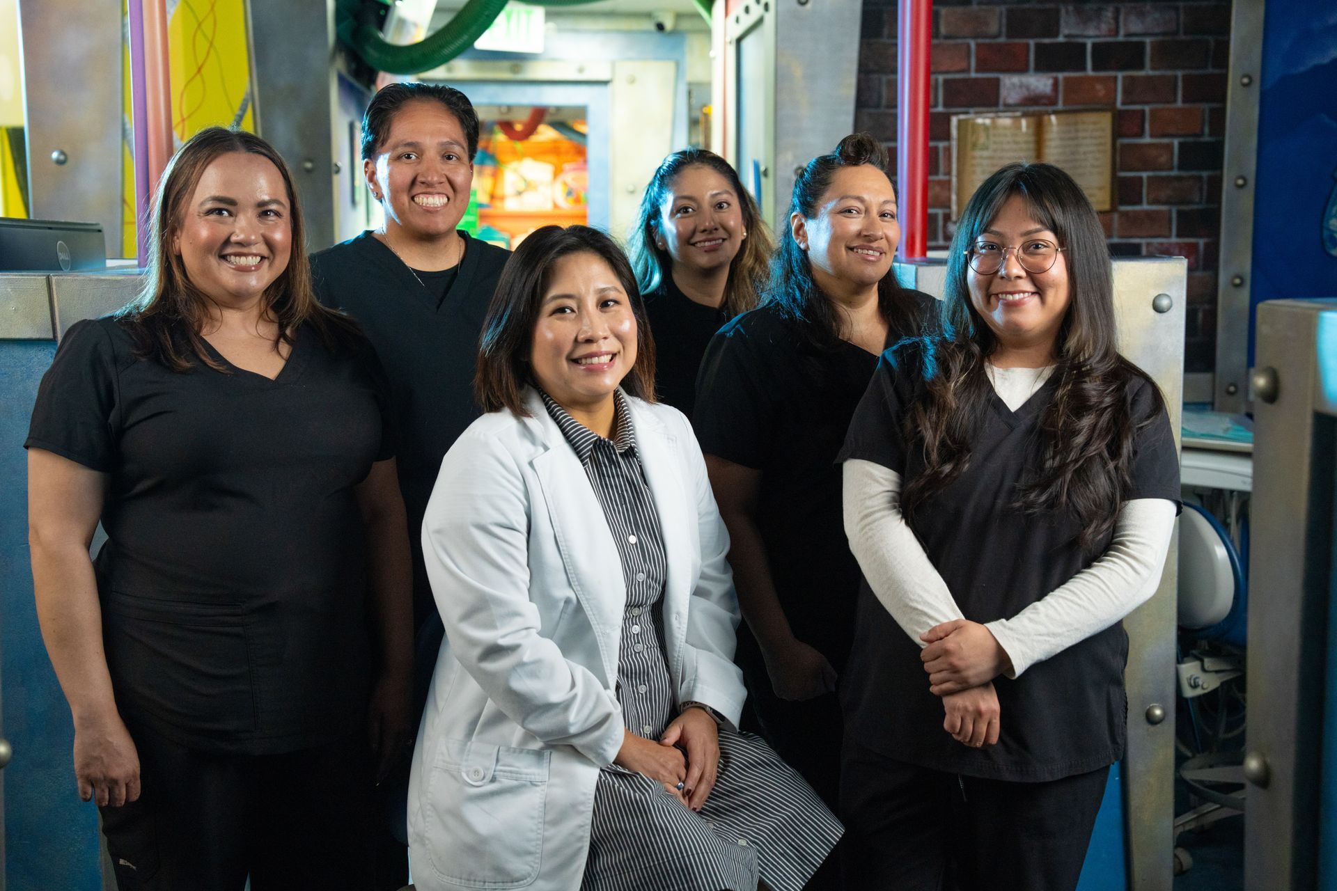 Six smiling coworkers standing in an office or workshop doorway, with one seated in front.