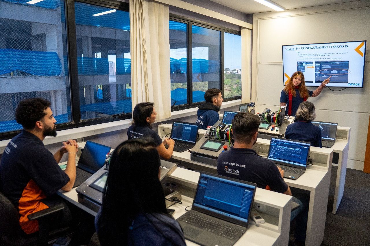 Uma mulher apresenta uma aula usando uma tela. Os alunos estão sentados em frente aos computadores em uma sala de aula moderna.