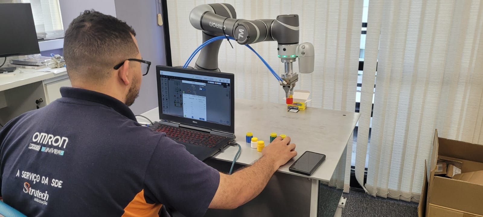 Uma pessoa programa um braço robótico com um laptop, que está interagindo com pequenos blocos sobre uma mesa.