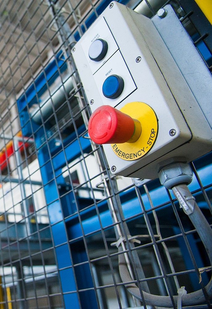 Botão vermelho de parada de emergência em um painel de controle, cercado por uma gaiola protetora azul e metálica.
