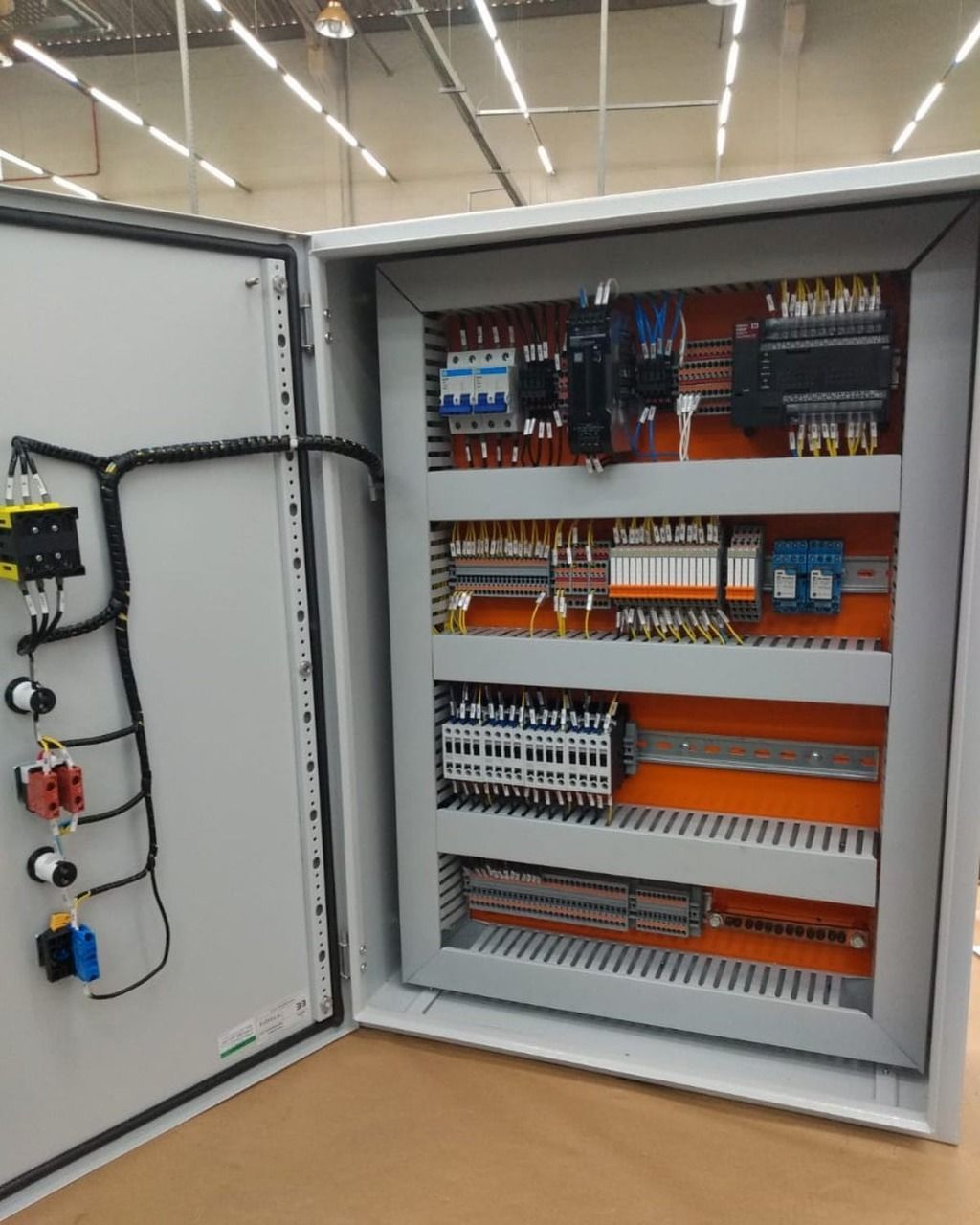 Painel de controle elétrico aberto com vários componentes sobre fundo laranja, fiação.