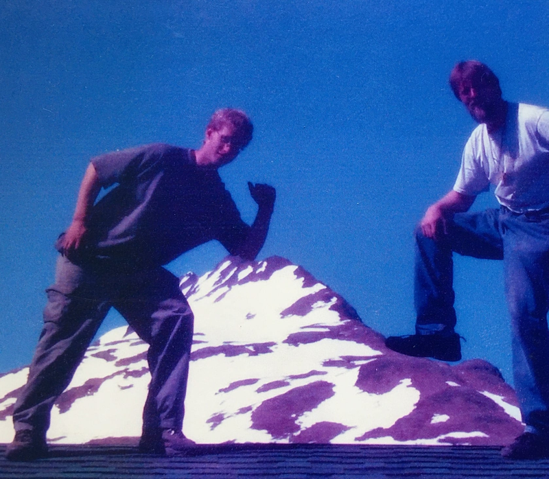 Two people pose in front of a snow-capped mountain against a clear blue sky.