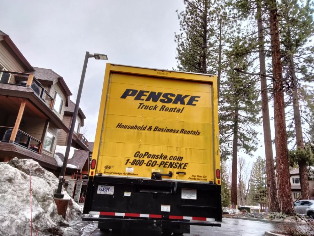 Yellow Penske truck backed into snowy driveway beside houses and tall pine trees