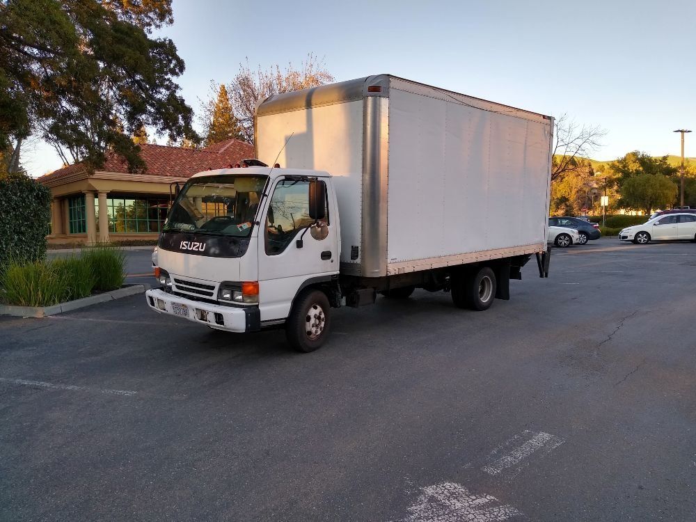 White box truck parked on a street at dusk, with trees, buildings, and other cars nearby.