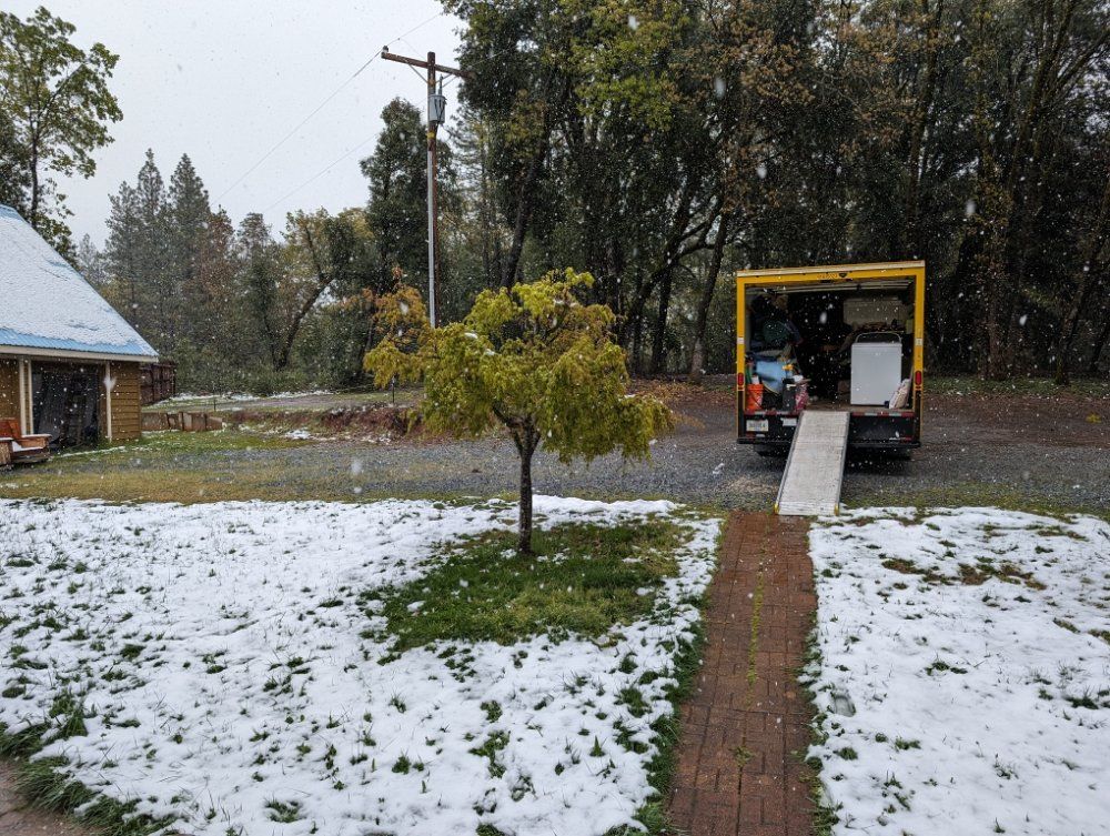 Snow-dusted yard with a tree and a truck loading ramp in light snowfall.