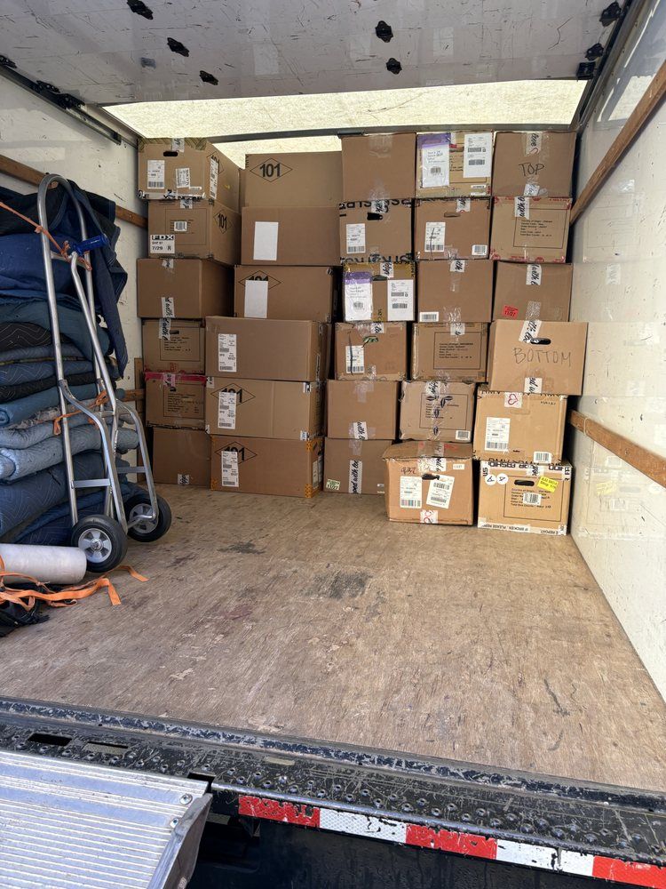 White moving truck with rear ramp open, revealing a large crate inside on a sunny lawn.
