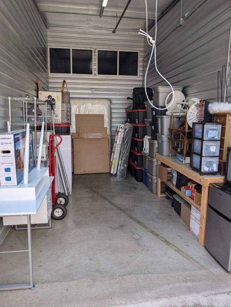Narrow storage room with shelves, boxes, tools, and stacked household items on both sides.