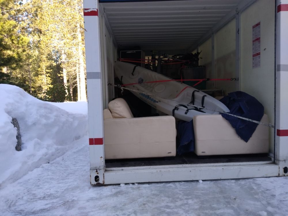 Open moving truck packed with a couch, bed frame, and boxes, parked beside snowy trees.