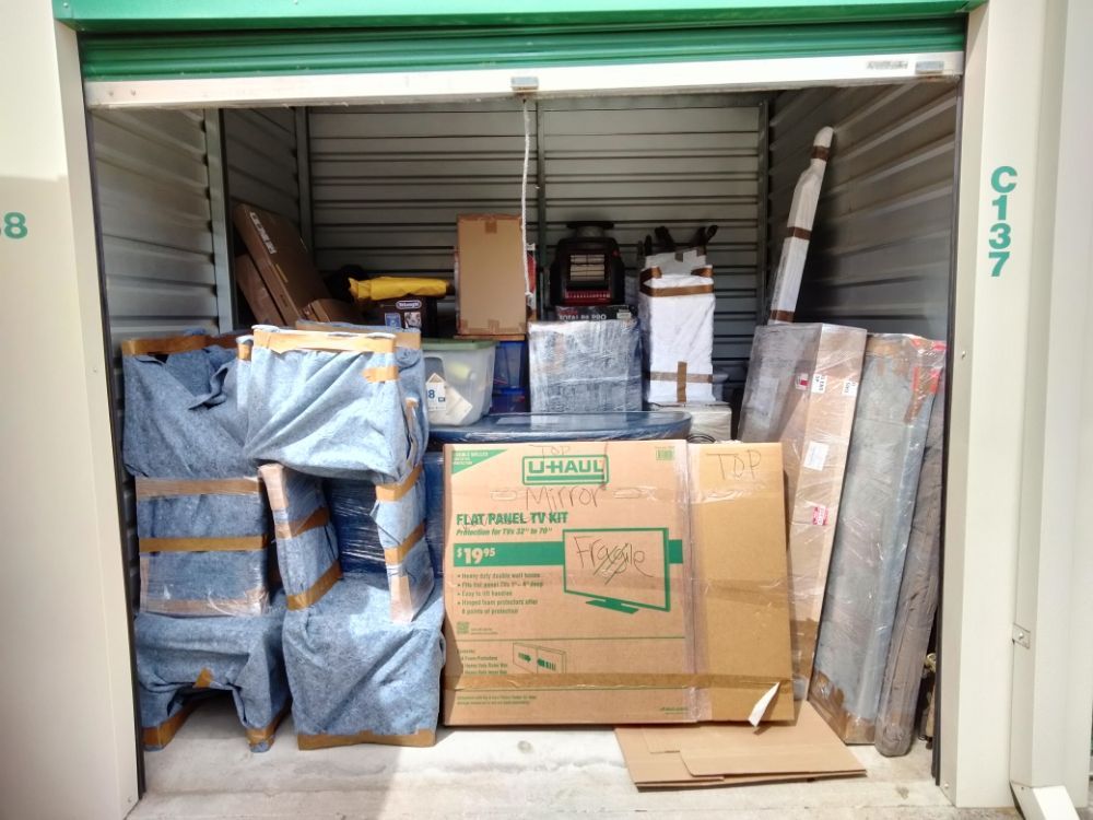 Storage unit packed with boxes, wrapped furniture, and stacked items under a partially open green door