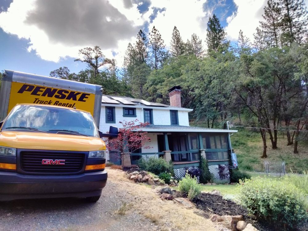 Yellow Penske moving truck parked beside a white house on a wooded hillside