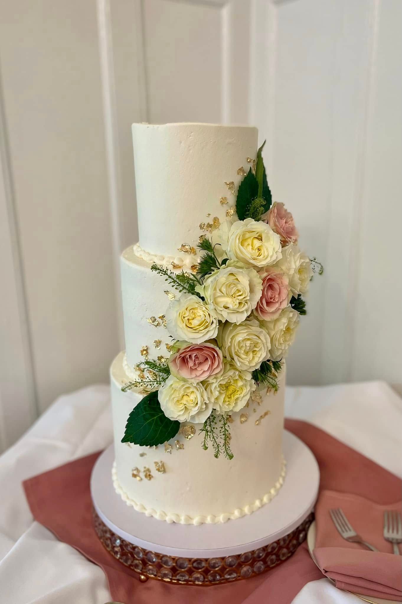 Three-tiered white cake with floral arrangement, resting on a pedestal and table with place setting.