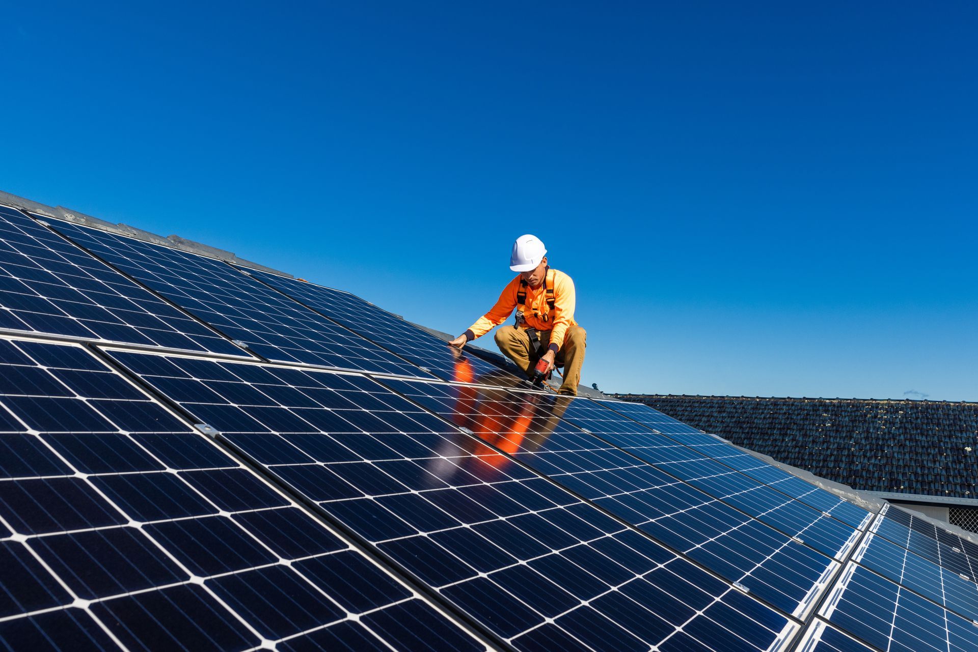 A worker in a white hard hat and yellow shirt inspects a large array of solar panels against a clear blue sky.