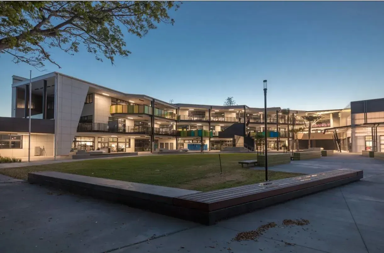 A modern, multi-story school building at twilight, overlooking a central grass courtyard with long wooden seating.