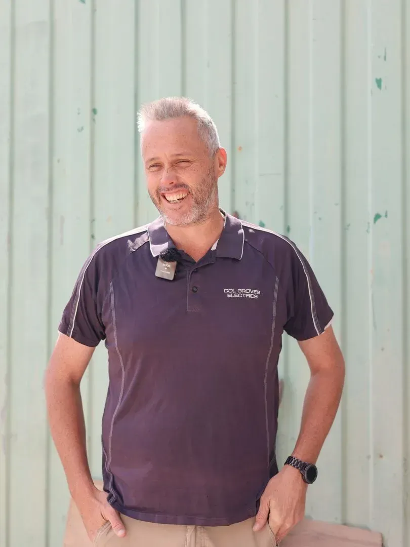 A smiling person wearing a dark polo shirt and a watch, standing against a light green paneled wall.