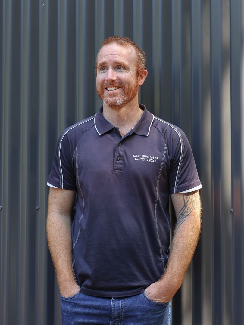 A person with a beard, wearing a dark blue polo shirt, stands with hands in pockets against a grey corrugated metal wall.