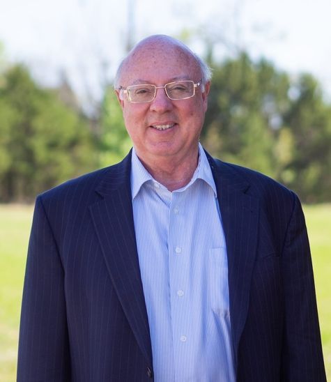 Smiling man (David Wilson) in a navy blazer and light blue shirt standing outdoors with blurred trees behind him