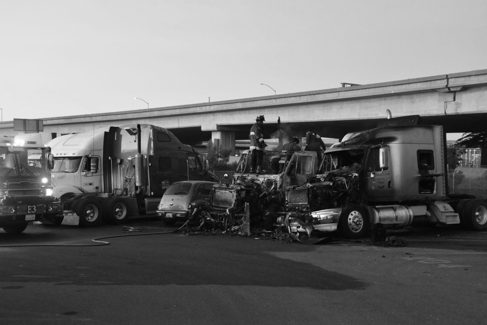 Two semi trucks badly damaged and tangled together on a roadside near an overpass.