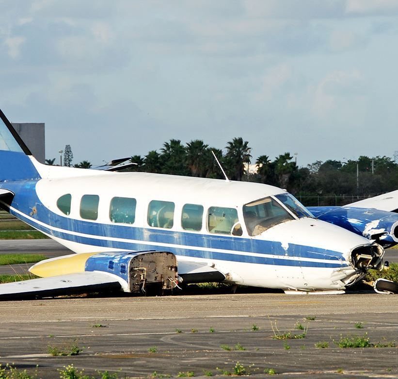 Damaged small airplane on runway, nose crumpled and wings broken, with blue and white livery