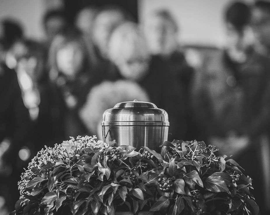 A silver urn resting on a flower-covered casket, with mourners blurred in the background.