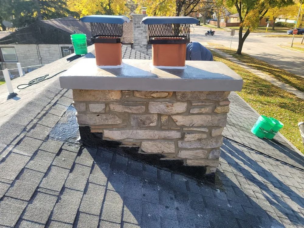 Chimney on a roof with two flue caps. The chimney is made of stone and the roof is asphalt shingle.