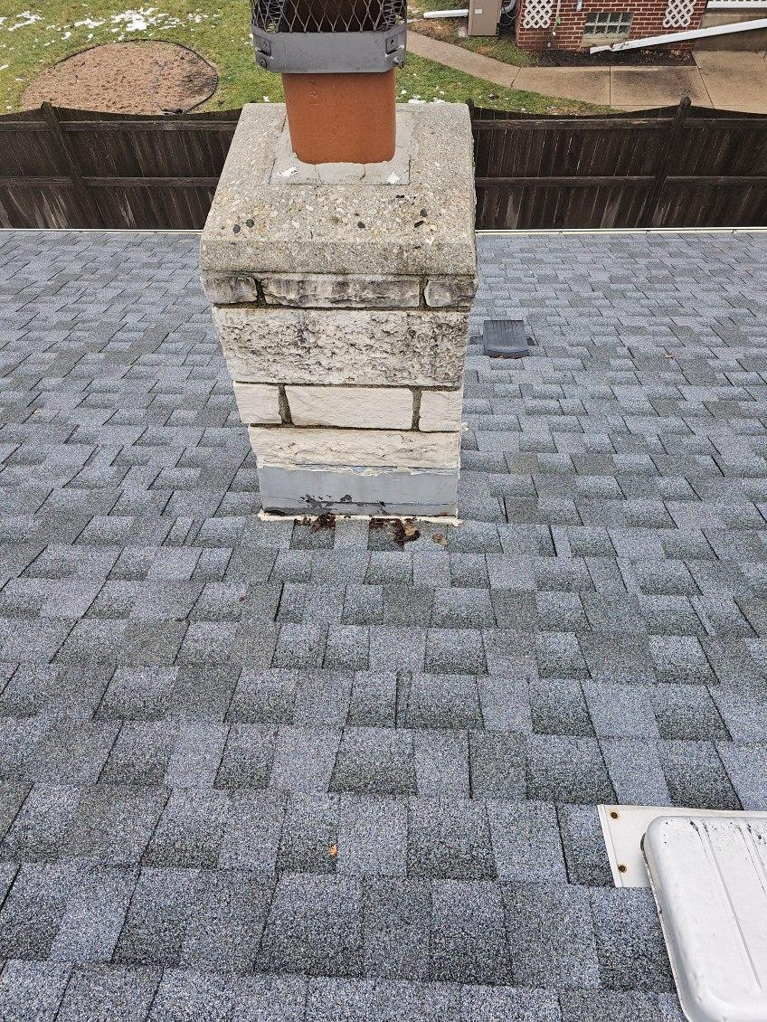 Chimney on gray shingled roof with a brown flue. Fence and snowy ground visible in background.