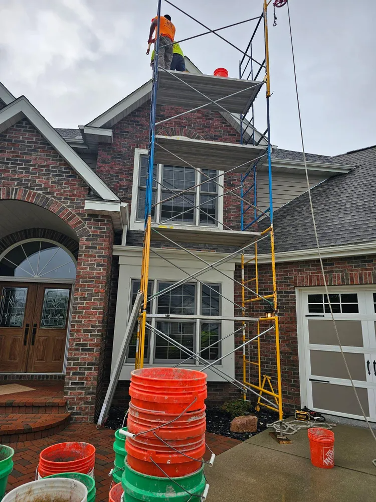 Workers on a tall scaffold next to a house with brick and a garage door, under a cloudy sky.