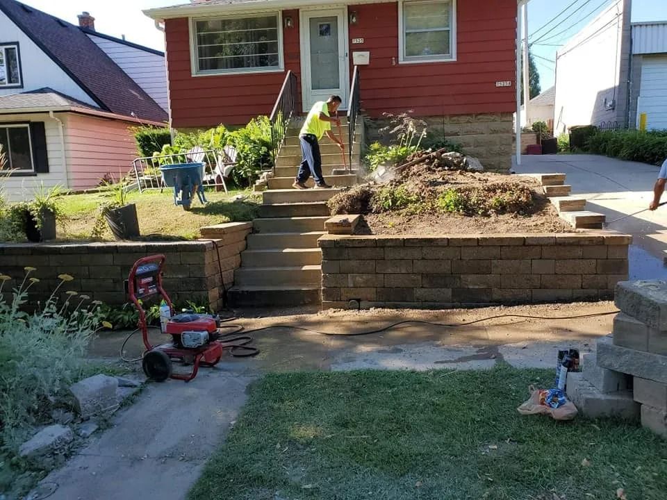 Man using a tool on steps in front of a red house with a retaining wall and grass.