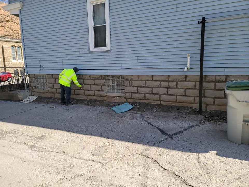 Person in neon vest inspecting brick wall base of blue building. Gray pavement in foreground.